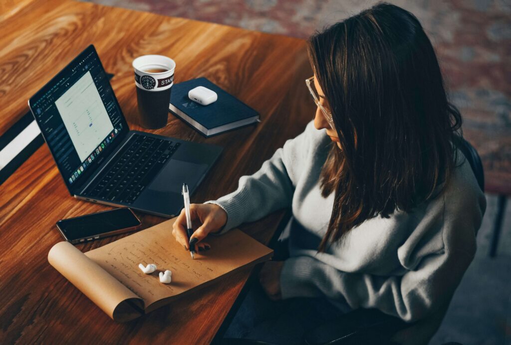 A woman is writing on a paper pad with her laptop next to it.