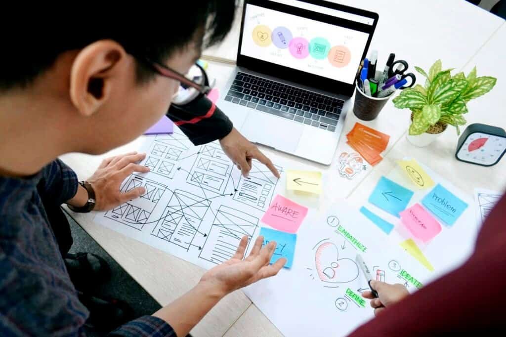 Designer sketching UI wireframes beside a laptop while learning UX design on a budget